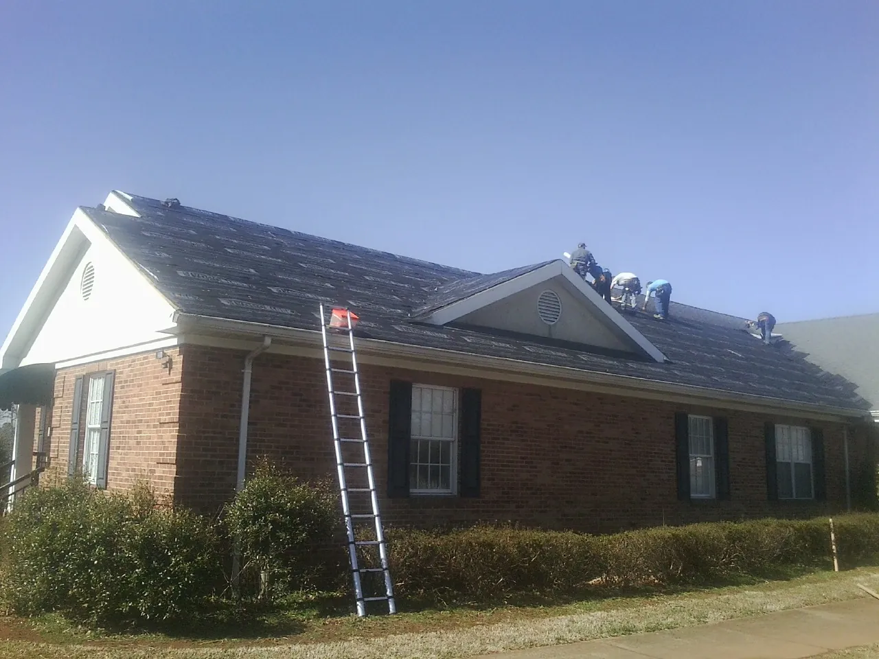 Crew tearing shingles off a brick ranch roof with a ladder set — Emergency Roof Repair work by our crew in Cataumet