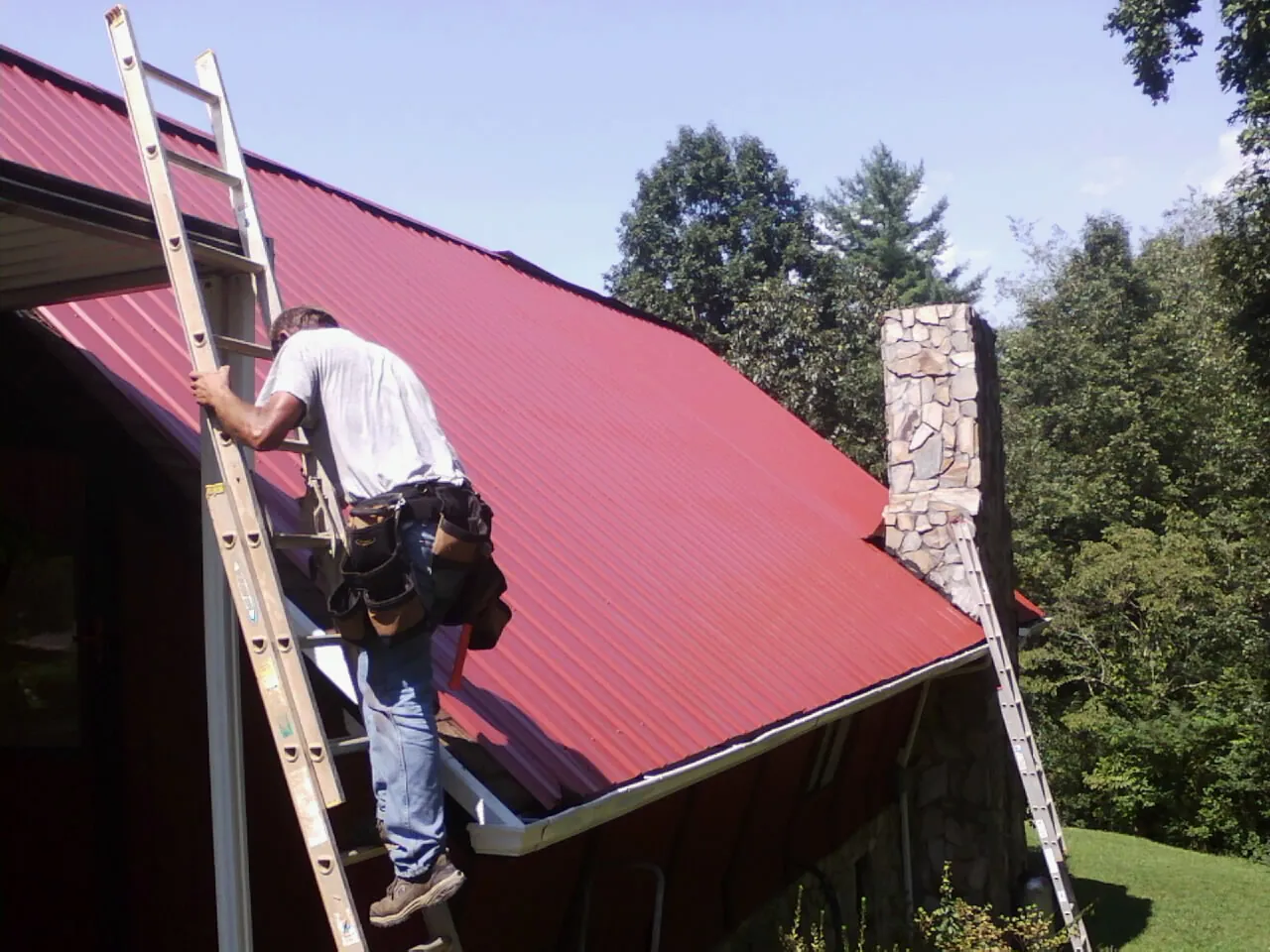 Roofer on a ladder beside a red metal roof and stone chimney — Soffit & Fascia Repair by Roofing Brothers in Cataumet